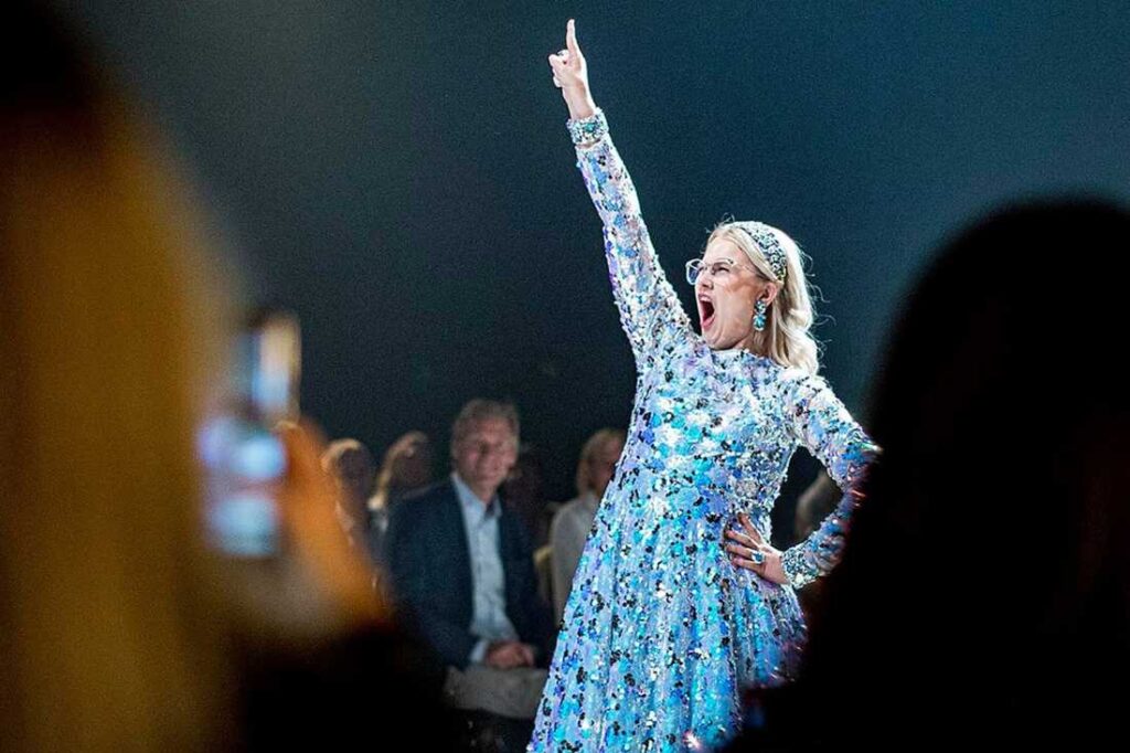 Emma är i bildens fokus iklädd en skimrande ljusblå klänning. Segervisst sträcker hon upp ena handen i luften och ser ut att ropa "JAAA!".