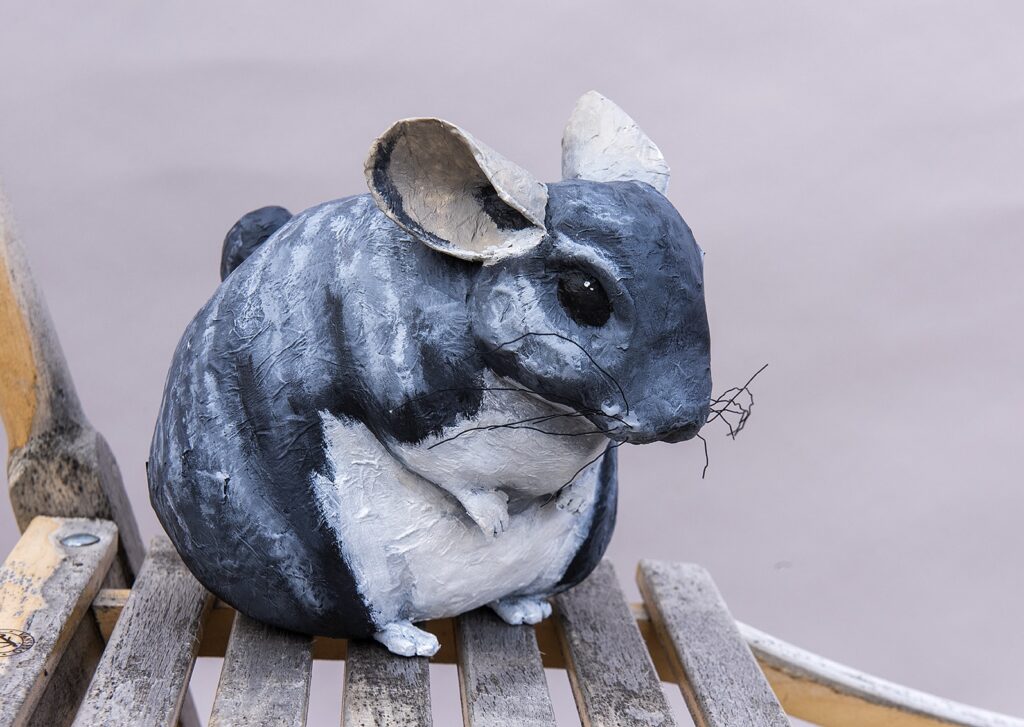 En skulptur av en chinchilla i papier-maché. Chinchillan sitter på marken och håller de små framtassarna framför sig. 