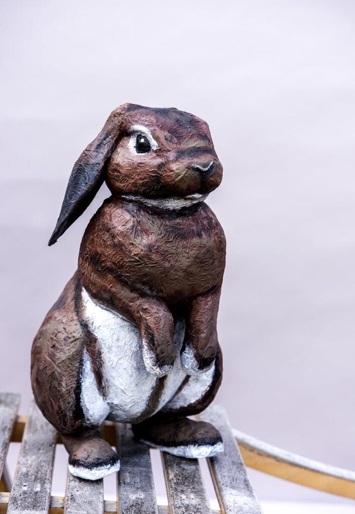 En skulptur i papier-maché som föreställer en kanin. Kaninen står upprätt på de stora baktassarna och den håller framtassarna framför sig över magen. 