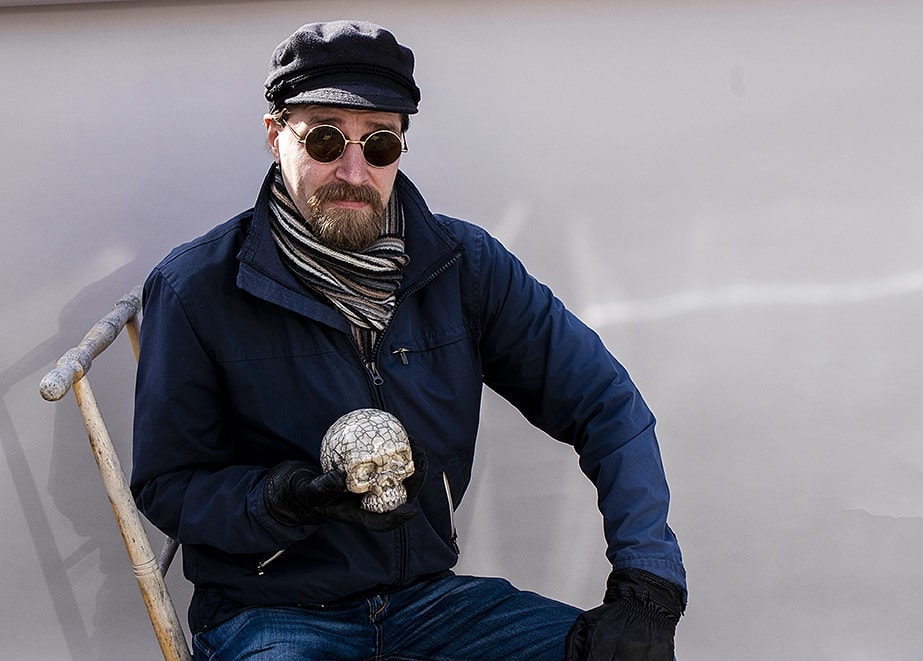 Mot en ljusgrå bakgrund utomhus sitter Robert på en spark. I sin högra hand håller han en raku-glaserad dödskalle.
