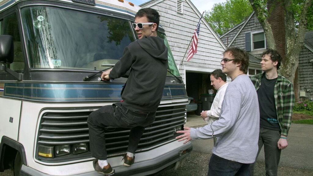 Noah står på kofångaren på husbilens front och håller i ett handtag. De tre andra killarna står runtomkring.