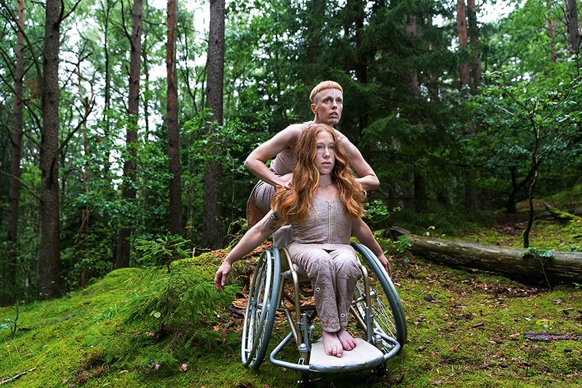 Omgivna av en grön och lummig skog på mossbeklädd mark ser vi Hannah och Felicia som sitter rakryggad i sin rullstol.