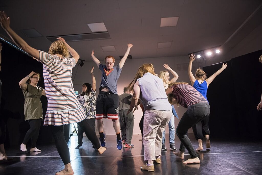 Nio personer i full rörelse på fotot i en danslokal. I centrum är en av de unga dansarna. Han är mitt i ett stort hopp med armarna uppsträcka i luften.