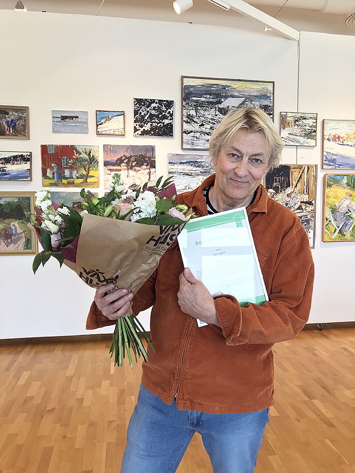 Lars Lerin står i utställningsmiljö med en stor bukett blommor och ett diplom. Lars tittar in i kameran och ler lätt.
