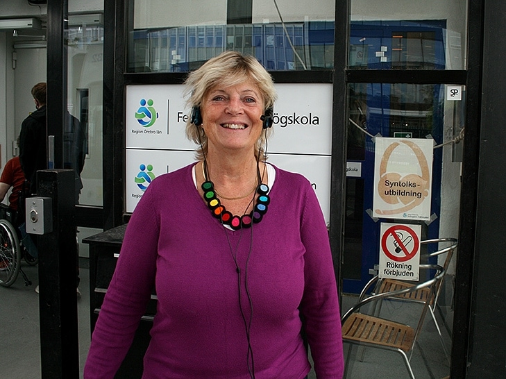 Porträtt på Lotta som står framför entrén till Fellingsbro Folkhögskola. Hon har på sig syntolkningsutrustning och ler glatt.