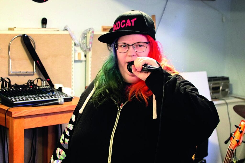 Sara i en replokal när hon övar på rap. Sara håller mikrofonen mot munnen och har en svart keps snett på huvudet där texten ”Foxocat” anas i rosa.