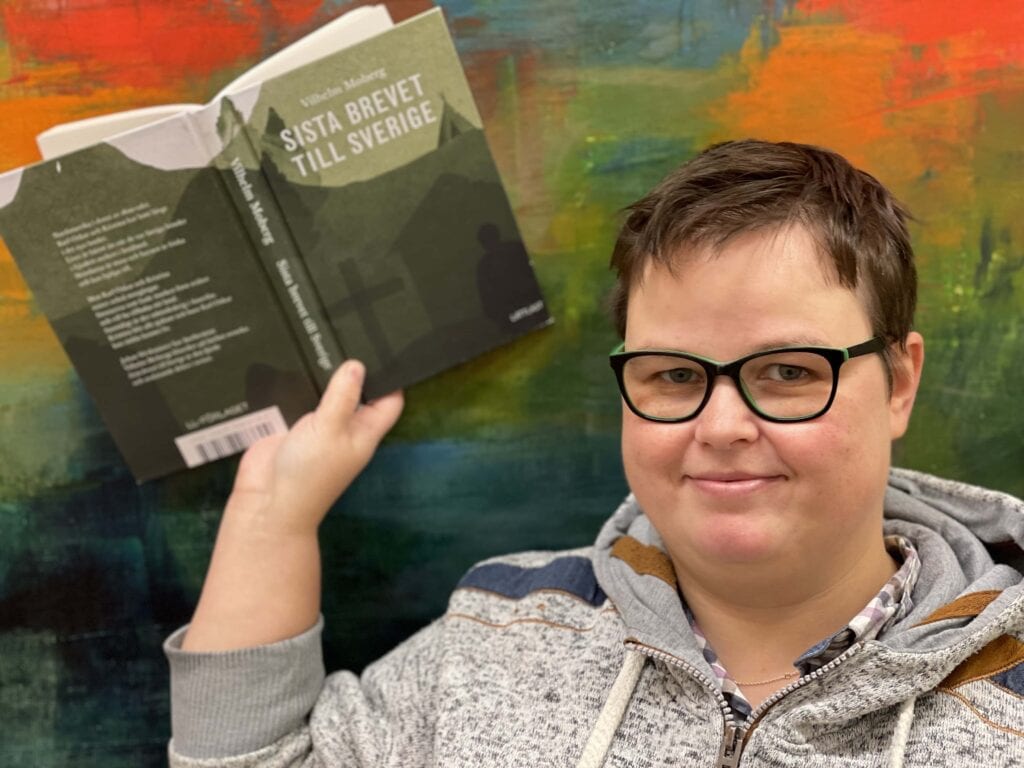Foto Kristina Stålnacke: Nina med boken ”Sista brevet till Sverige”.
Nina som håller den uppslagna boken i sin hand, så både framsidan och baksidan syns. Hon står mot en abstrakt färgad bakgrund i rött, orange, blått och grönt.