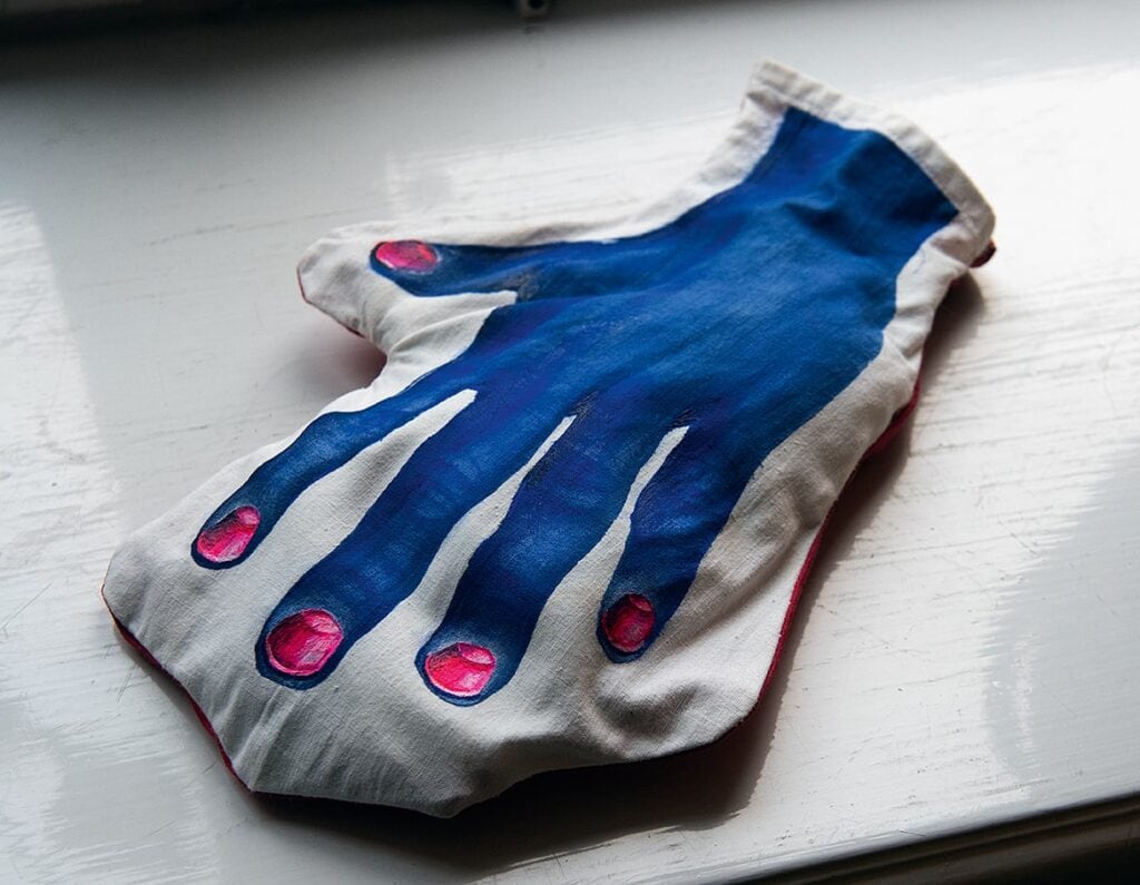 Vetevärmare tillverkad av vitt tyg och fylld av vetekärnor. Den har formen av en tumvante med en bild av en stor blå hand med rosa naglar tryckt ovanpå.