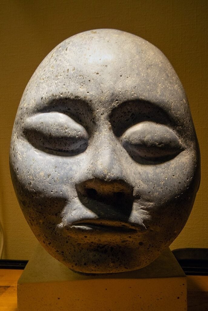 En mörkgrå skulptur föreställande ett ansikte med en form som är oval och placerad direkt på en fyrkantig ljusare sockel. Pannan är hög, Ögonhålorna är stora men ögonen är stängda, näsan är liten och nätt, kindknotorna är höga den raka munnen är stängd och de smala tunna läpparna putar utåt.
