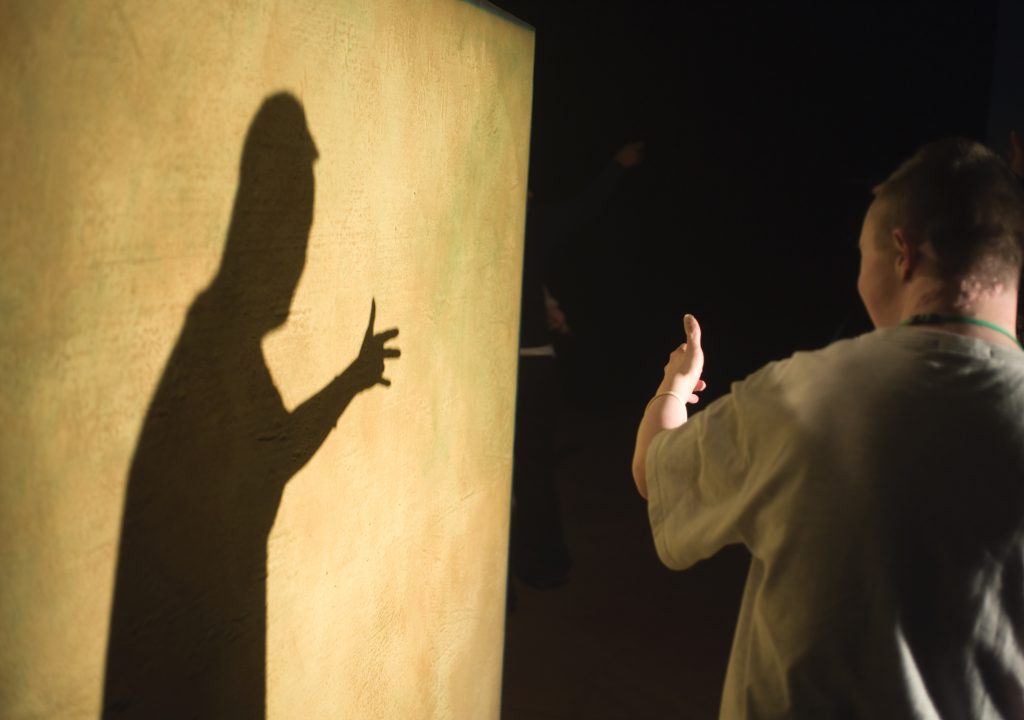 Foto: Svenska Mosaikteatern på utbildning hos Mind the Gap i England.
En ung kille i t-shirt i en mörk omgivning som står med ryggen mot oss och betraktar sin skugga mot en ljus vägg.
