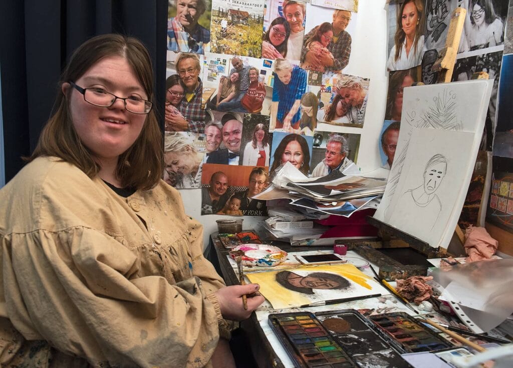 Johanna sitter vid sin arbetsplats med en pensel i handen, omgiven foton på olika personer.
