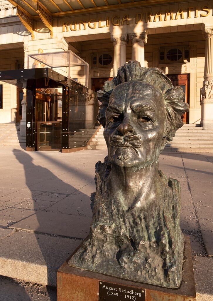 En bronsbyst på August Strindberg vid Dramaten, där solen kastar en lång skugga av honom som pekar mot hissen vid huvudentrén.