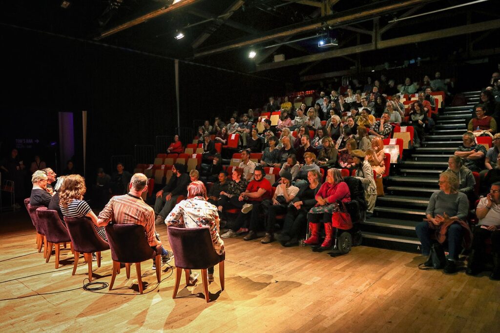 Ett sittande panelsamtal pågår på scengolvet och publiken sitter och lyssnar på talarna.