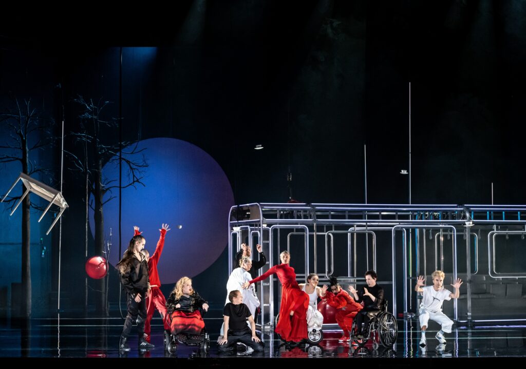 En scenbild med hela ensemblen på scen i rörelse. De är klädda i svart, vitt och rött. Bakgrunden är svart och månen syns. Konturerna av en tunnelbana i stålrör syns också.