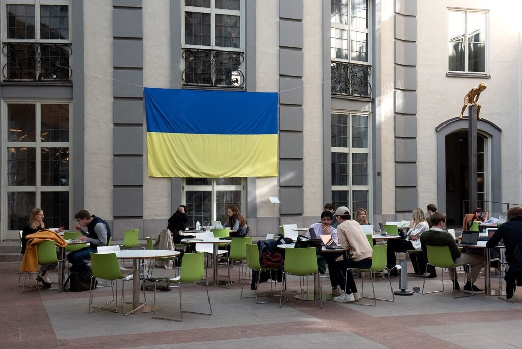 På en inomhusfasad hänger den ukrainska flaggan uppspänd i mitten på bilden. Till höger om flaggan en förgylld staty som ser ut att hoppa bock över en några meter hög pelare. På golvytan framför konstverken är flertalet bord med elever.