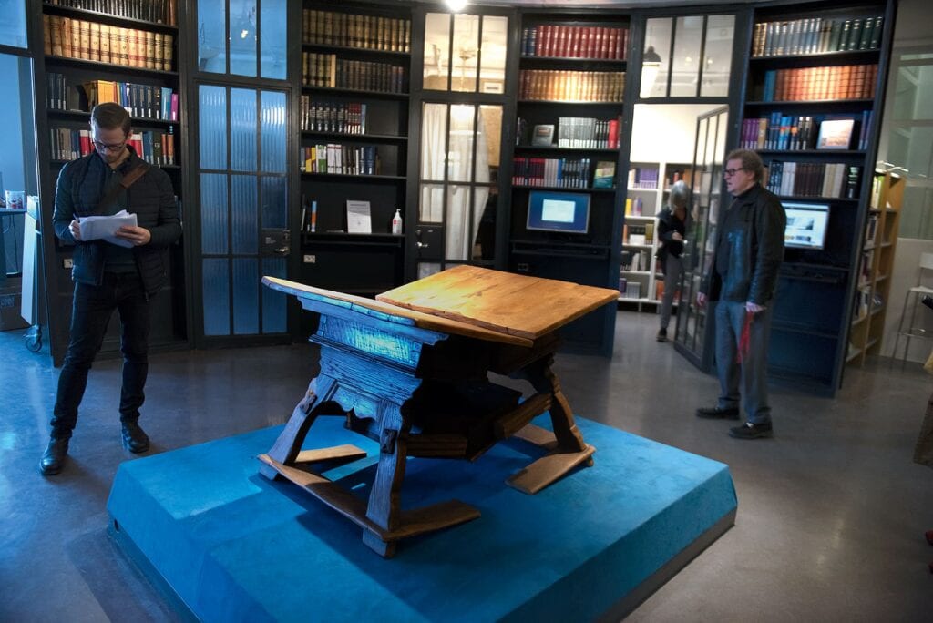 I mitten av ett slags biblioteksrum, ett lågt podium med en knäckt bänkskiva på en gedigen träkonstruktion.