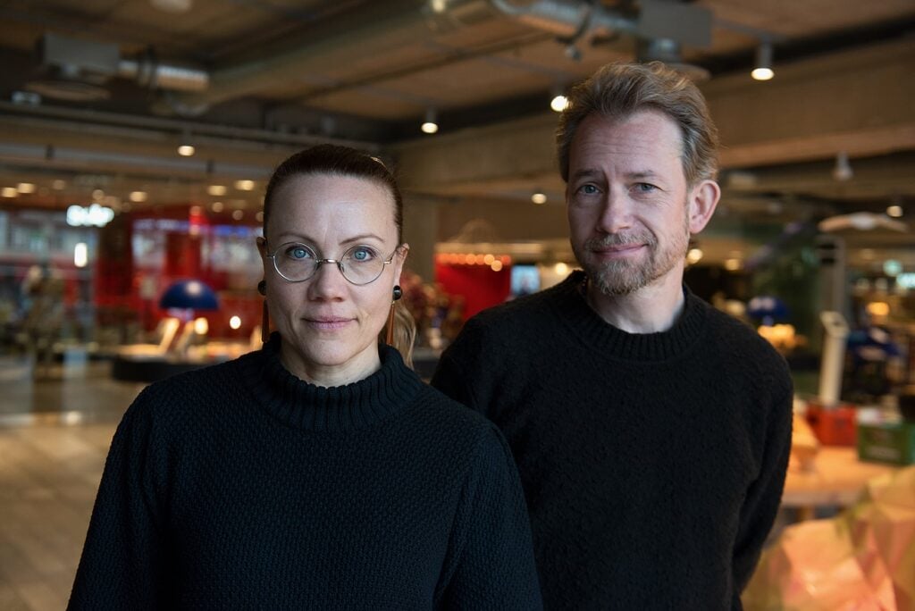 Porträttbild av Jenny Åhsberger och Po Hagström som står i en stor industrilokal med röda väggar och ventilationsrör i taket.