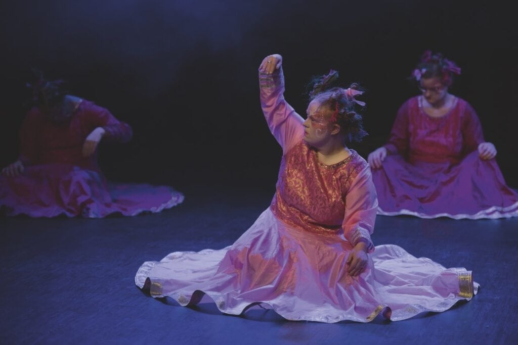 Tre kvinnliga dansare i rosa klänningar sitter på golvet. Fokus på en dansare som höjer sin arm och ser allvarlig ut.