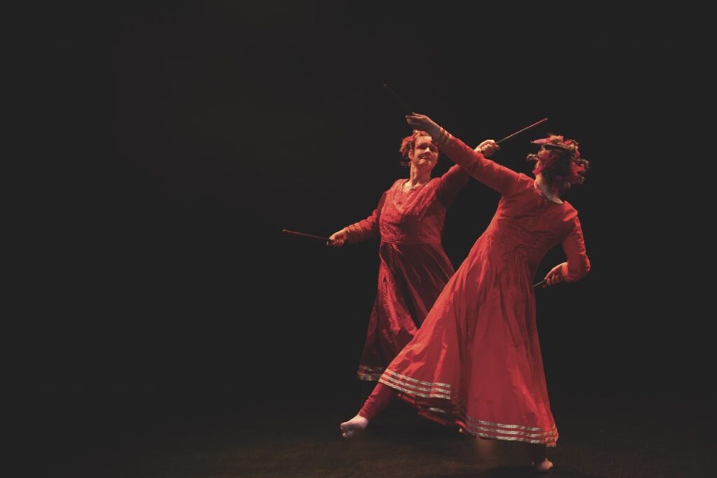 Två kvinnliga dansare i röda klänningar med korta käppar i händerna, möter varandra som de dansar och fäktas.