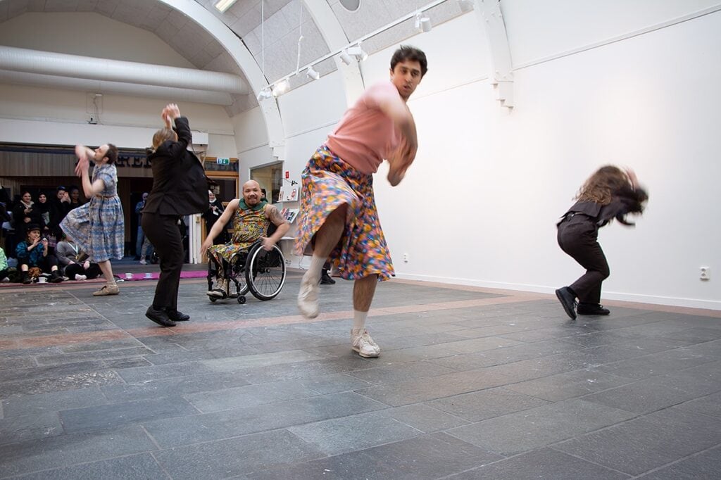 Fem dansare i full fart på ett stenbelagt golv i inomhusmiljö. En av dansarna sitter i rullstol. Dansarna rör sig åt olika hålla i svepande rörelser.