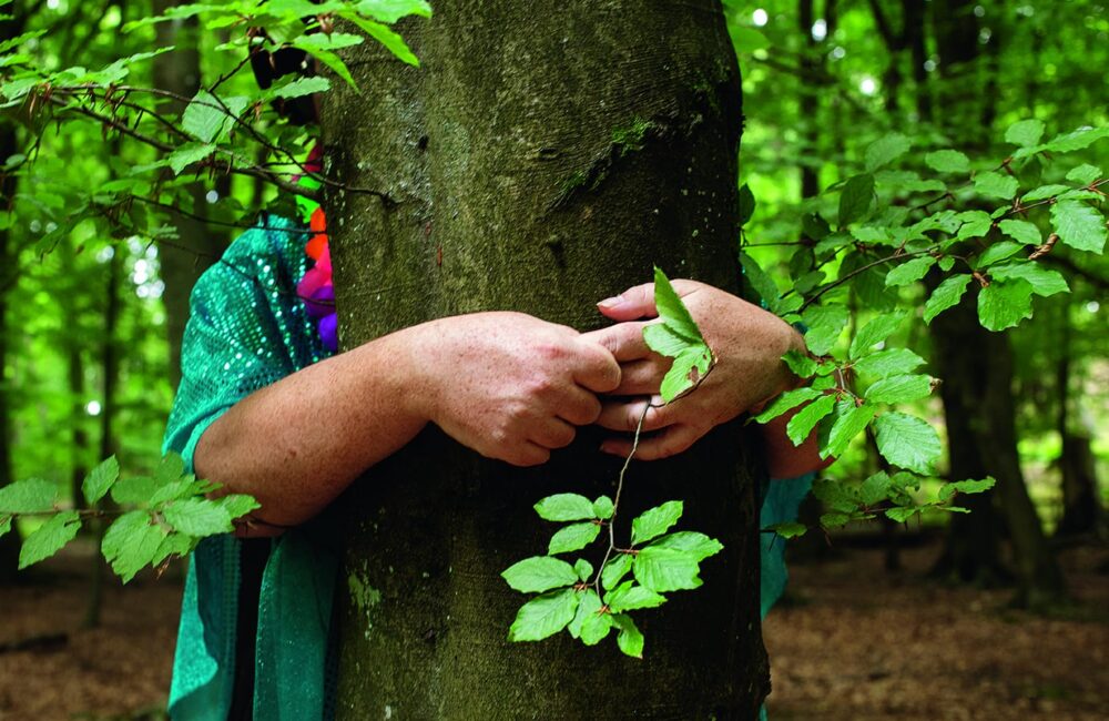 I en grön lövskog syns två armar krama en trädstam. med en gestalt som syns bakom trädet.