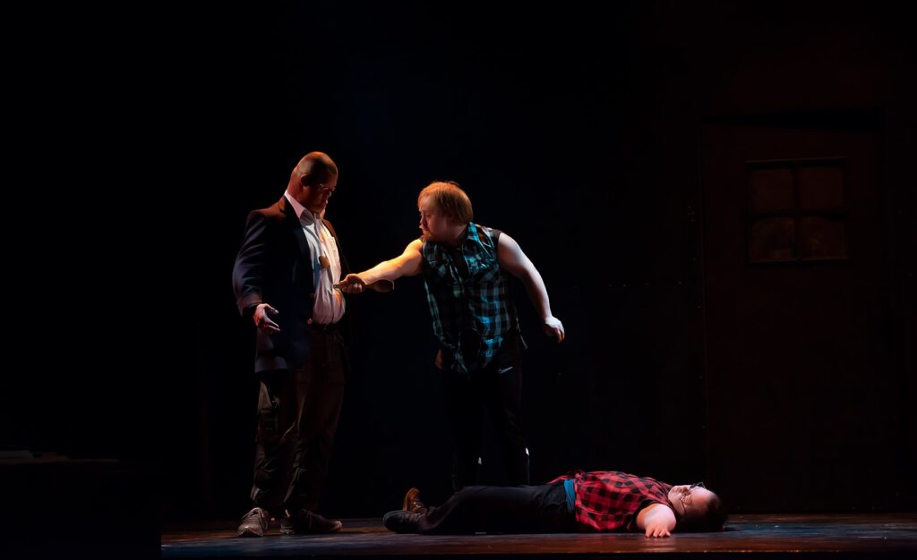 En scenbild med tre personer mot en svart bakgrund. En ung kvinna ser ut att ligga död på golvet, en ung man sticker skaftet på en träslev i magen på en annan man.