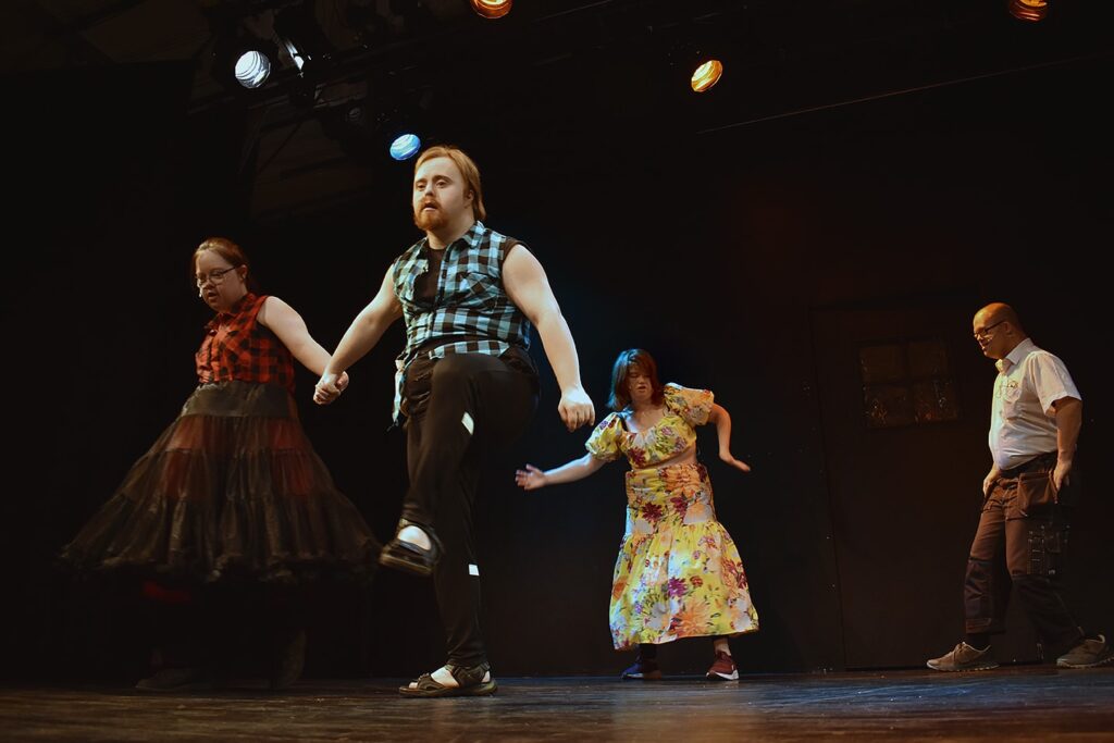 Scenbild på fyra personer med svart bakgrund. I den främre delen av på scenen står två deltagare i kursen, en man och kvinna, och håller varandra i händerna. I bakgrund två personer i rörelse.