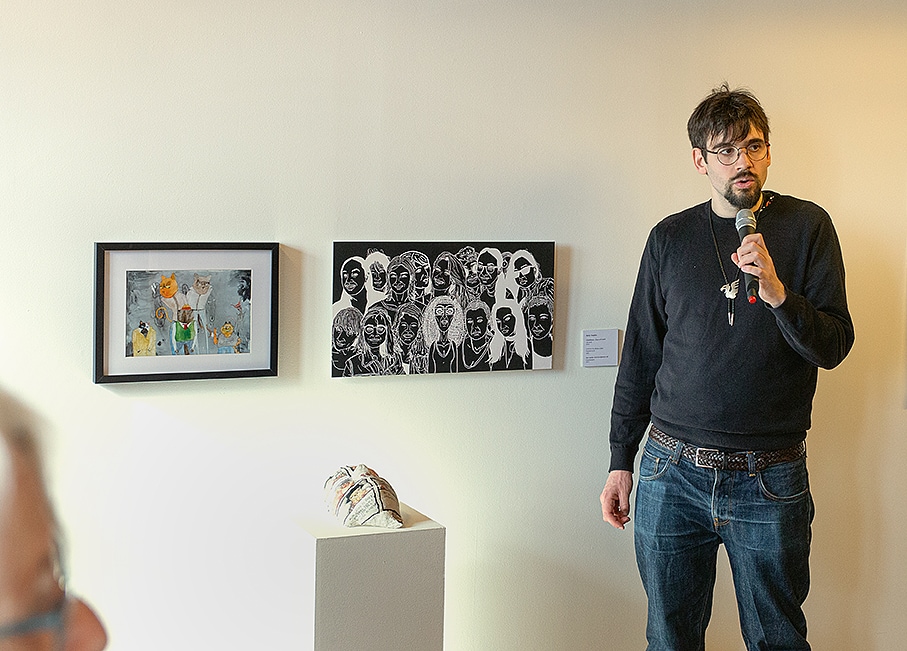 Felix Swahn står med en mikrofon i handen bredvid sina konstverk och pratar. Till vänster om honom hänger två tavlor på väggen och på en piedestal framför tavlorna ligger en skulptur med formen av ett ansikte.