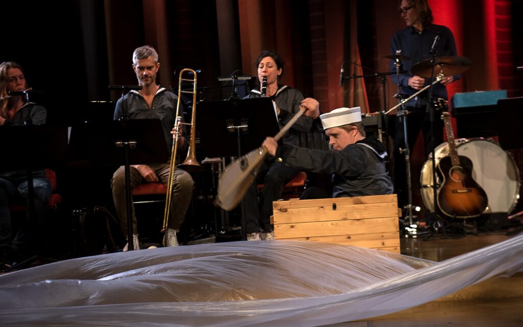Scenbild med en ung man med Downs syndrom och sjömanskläder sitter i en trä-lår och ror, bakom honom syns fyra musiker.