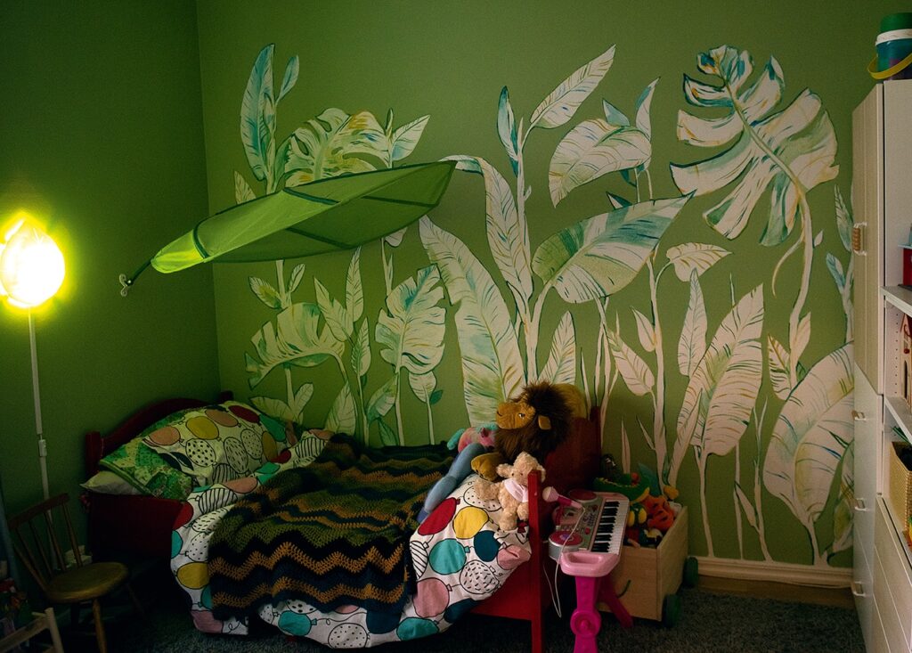 Ett fotografi av en väggmålning i ett i övrigt grönmålat rum med en kort låg säng med plyschdjur som ligger vid fotänden. Den gröna väggen är täckt av målade stora växter. Bladen är ljusa, nästan vita med skuggor i olika gröna nyanser.