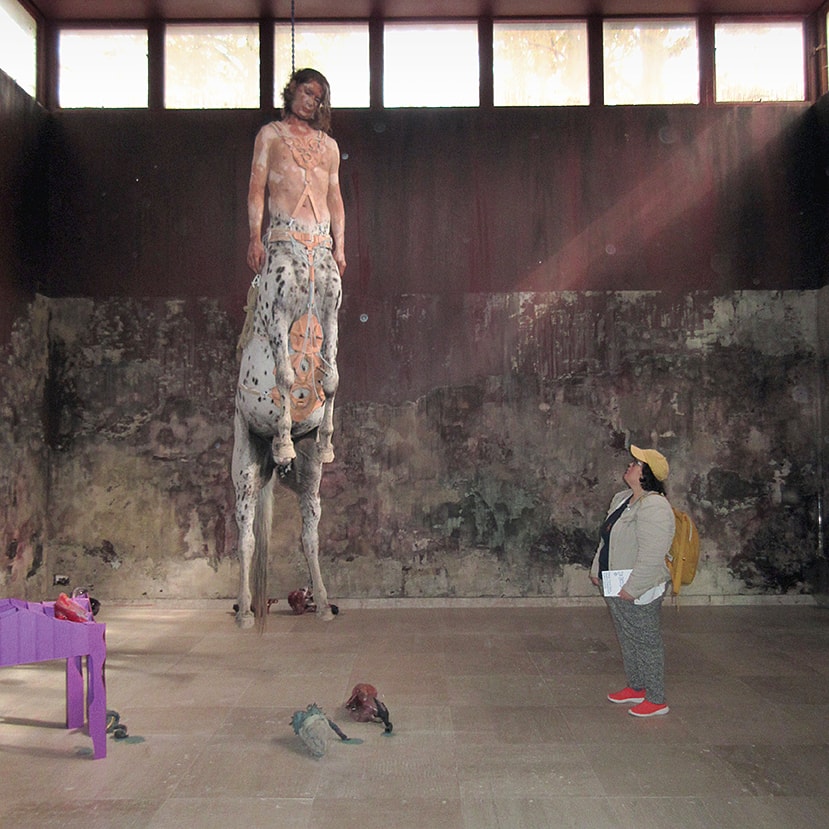 Skulpturinstallation med en slags död kentaur som tycks hänga i en strypsnara. Hanna står bredvid kentauren på golvet och kollar upp på den hängande gestalten.