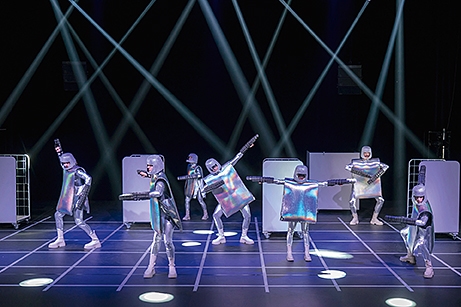 Scenbild med sju personer i silvriga kvadratiska kläder som ger ett robotliknande intryck tycks utföra en dans.