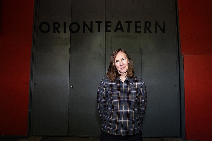Ett fotografi på Jonna som står framför en mörk, grafitfärgad vikvägg. På väggen står det Orionteatern i svarta versaler - teaterns logotyp.