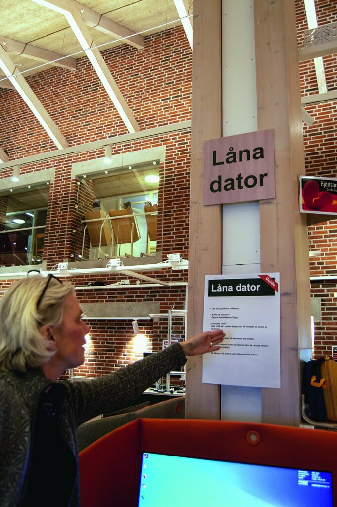 Vid en pelare sitter en skylt om att ”Låna dator” och under en lapp med instruktioner som bibliotekarie Lisa pekar på. 