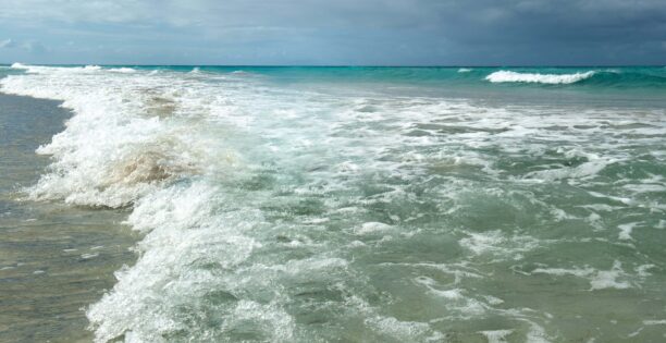 Ett vidsträckt turkosfärgat hav med vågor som slår upp mot en sandstrand.