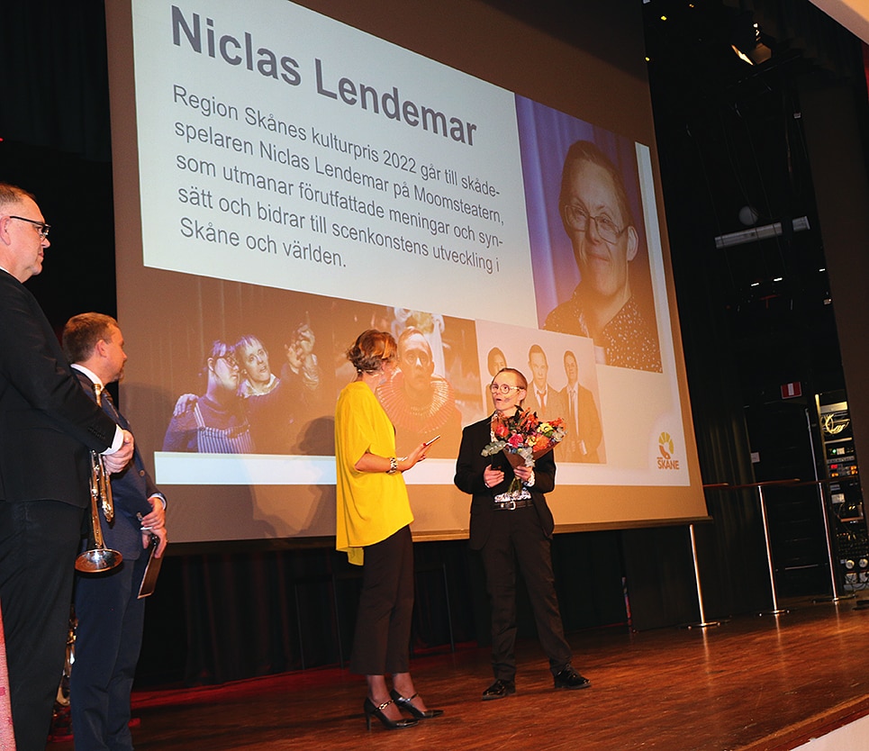 Prisutdelning på en scen i Eslövs kultur och medborgarhus den 30 november 2022. Niclas Lendemar tar emot priset av en kvinna med uppsatt hår och starkt gul blus.