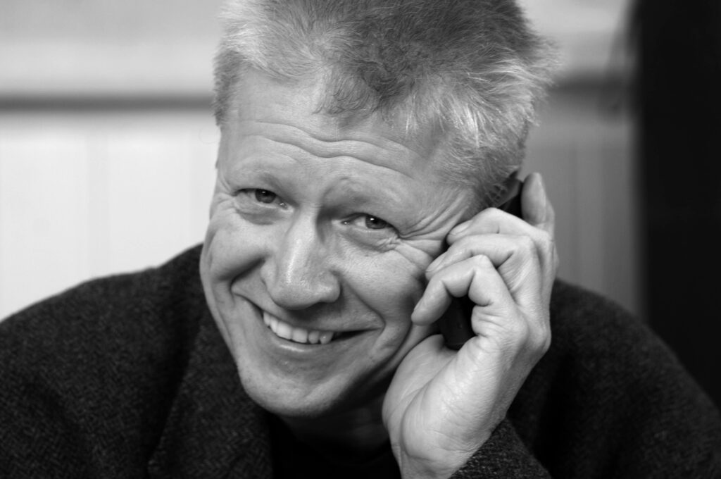 Svartvitt porträtt på Martin som har en telefon tryckt mot örat och leende tittar in i kameran.