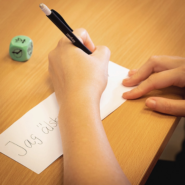 Närbild på en hand som skriver med blyerts på en lapp som ligger på ett bord. 