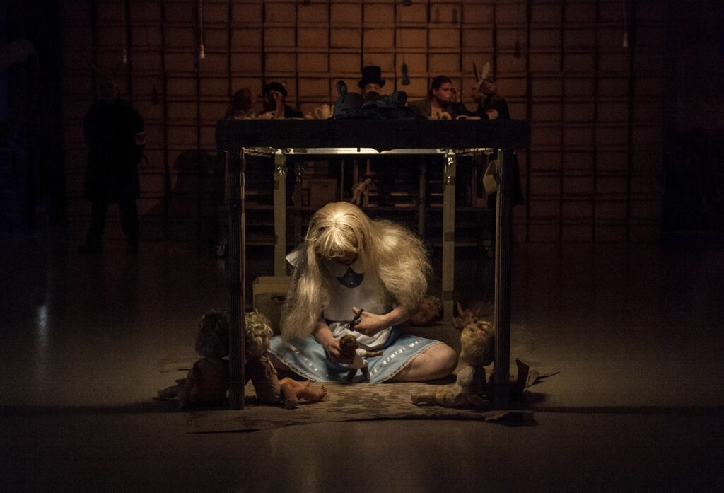 Foto från teaterföreställningen ”Wonderland” på Moomsteatern 2019. På en mörk scen i en fyrkantig metallbur sitter en person med långt blont hår och lugg på en tunn fäll.
