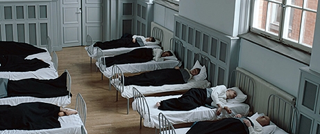 Flera sängar på rad utmed en vägg med personer som ligger i dem i en stor och ljus sal. 