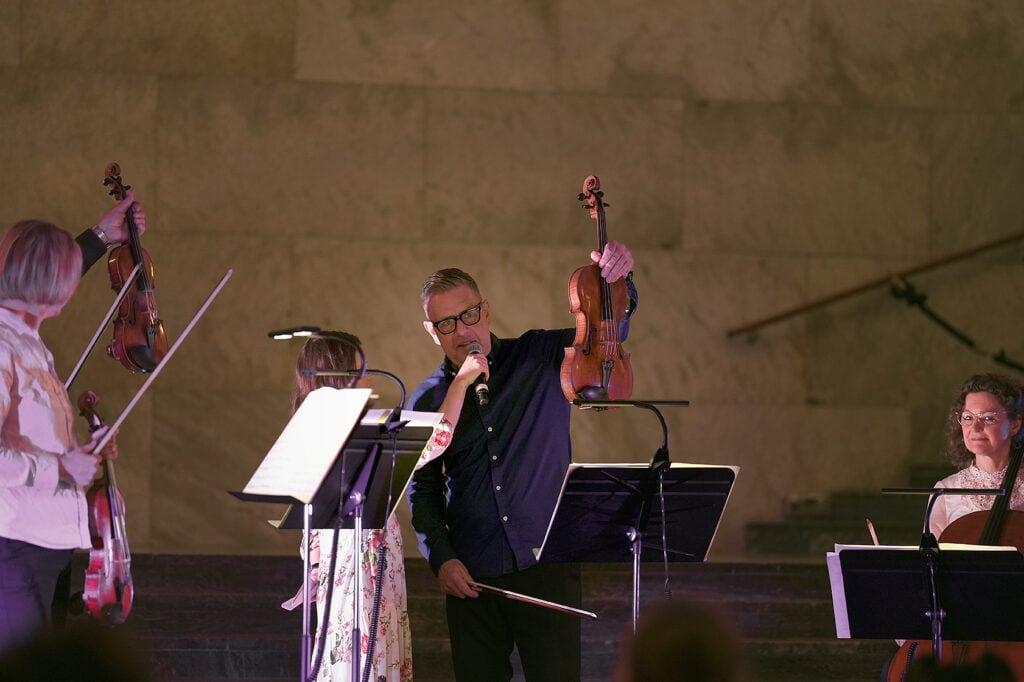 Scenbild med Lars som håller upp sin altfiol i luften och pratar i en mikrofon som en kvinna i blommig klänning håller fram.