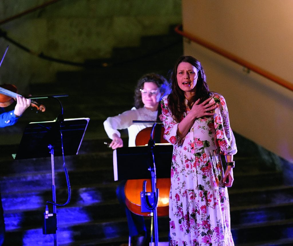 Scenbild med fokus på en kvinna i blommig klänning sjunger har lagt vänsterhanden över sitt bröst och ser fokuserad ut. Bakom anas en cellist som spelar.