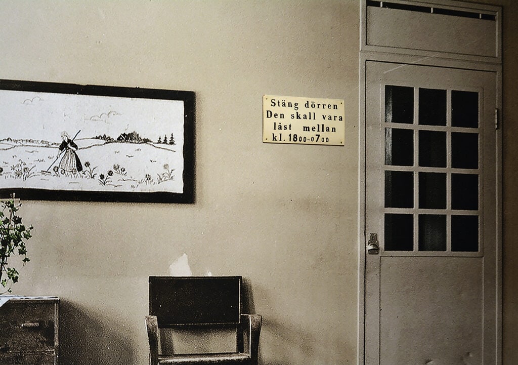 Ett gulnat svartvitt foto med en kal ljus vägg med en stängd dörr och en skylt i gammal stil som säger när dörren ska vara stängd.