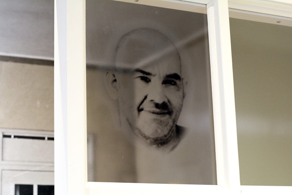 På ett mindre glasfönster sitter ett halvtransparent foto som föreställer en man med rakad skalle, och kraftiga markerade ögonbryn. Han ler lätt. 