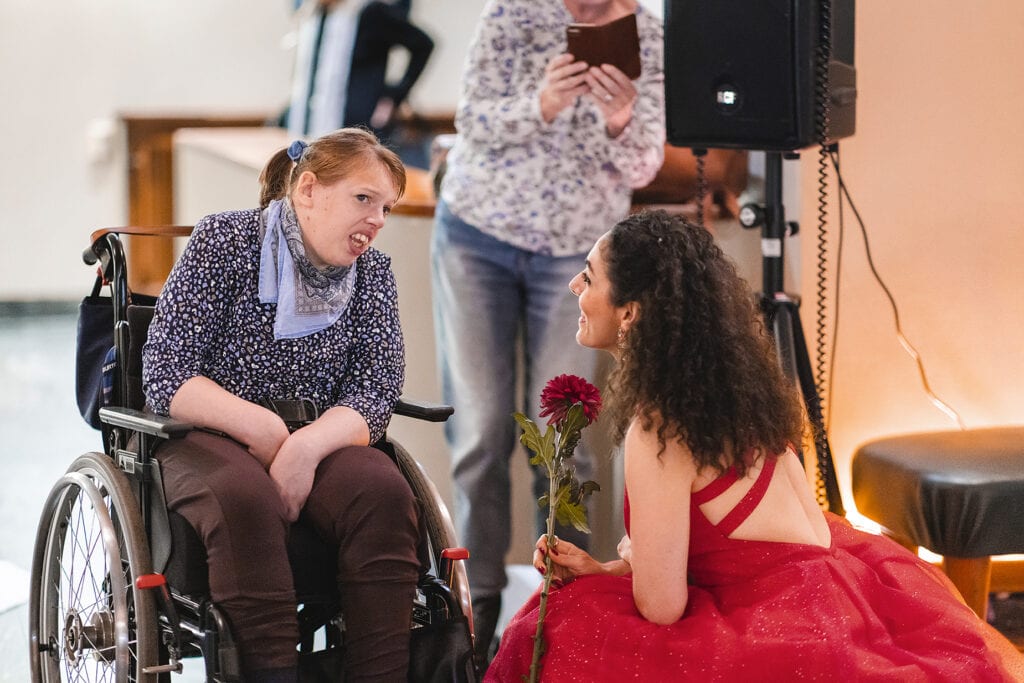 Den rödklädda sångerskan sitter med en blomma i handen och pratar med en ung tjej som sitter i rullstol.