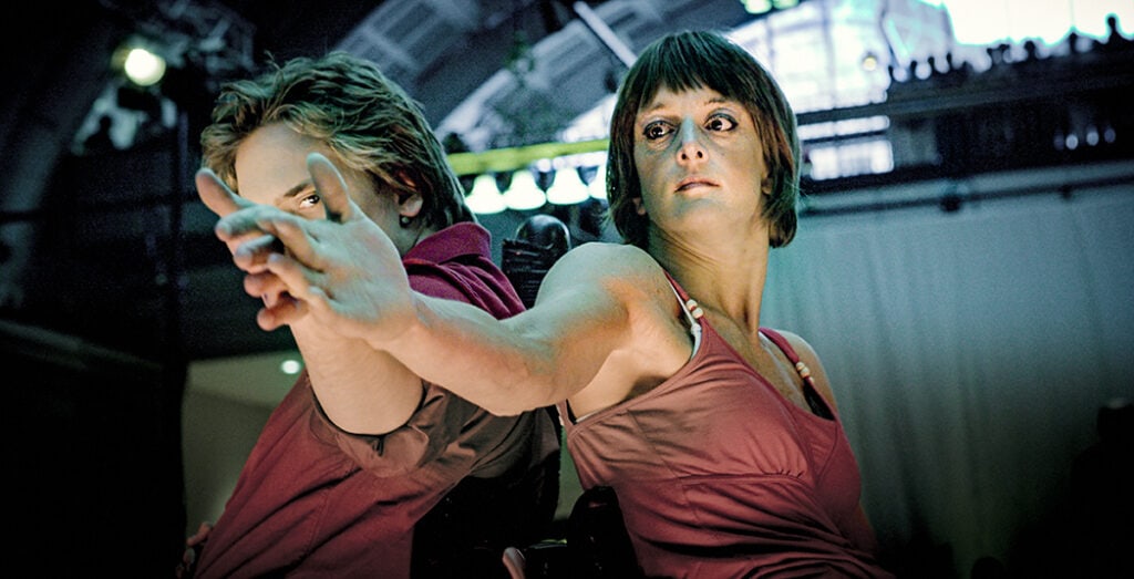 Scenbild på två dansare med ryggarna mot varandra som håller ut armarna rakt ut så deras handryggar möts. Fokus i bilden är på de utsträcka händerna som mötts.