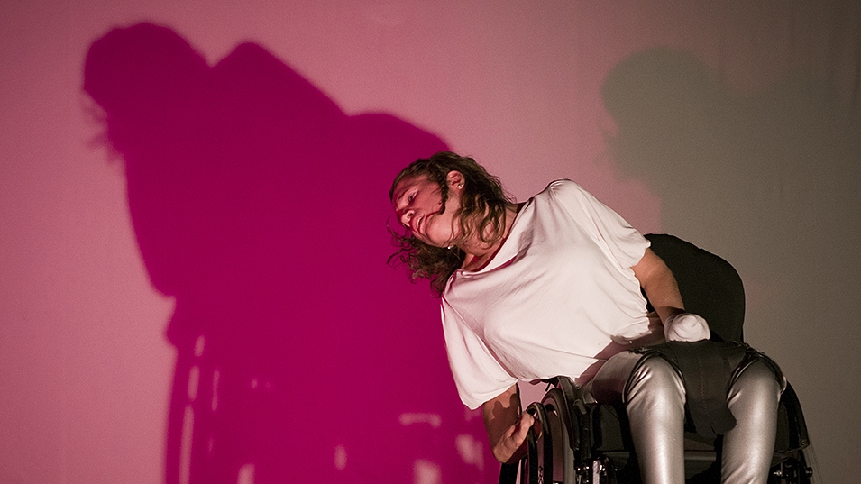 Scenbild på Karin som lutar sig snett bakåt och åt sidan i sin rullstol i en dramatisk posé med håret slängande runt ansiktet. Hennes skugga är skarpt rödrosa mot den mjukt rosa bakgrunden.