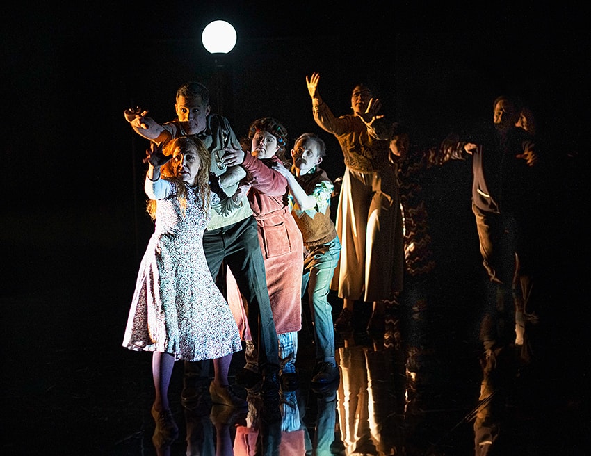 Scenbild ur föreställningen ’Under fullmånen’. En blandad grupp om åtta personer står på rad bakom varandra. Niclas, som står bland dem, har pressade byxor, kortärmad skjorta och brun väst.