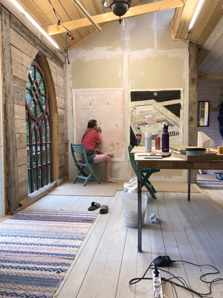 Toffe sitter på en stol i en ateljé och målar på ett konstverk. Rummet är högt och ljust. Ett stort kyrkfönster med träinfattning går från golv till tak.