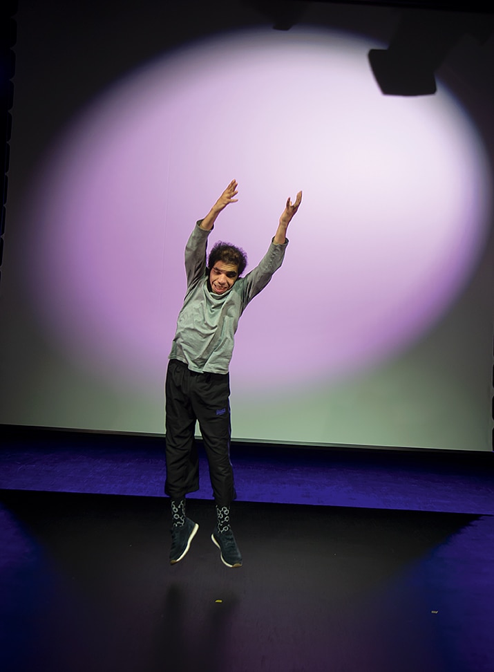 Den mörkhyade dansaren DJ befinner sig i luften i ett hopp med händerna högt sträcka över sig på en scen.